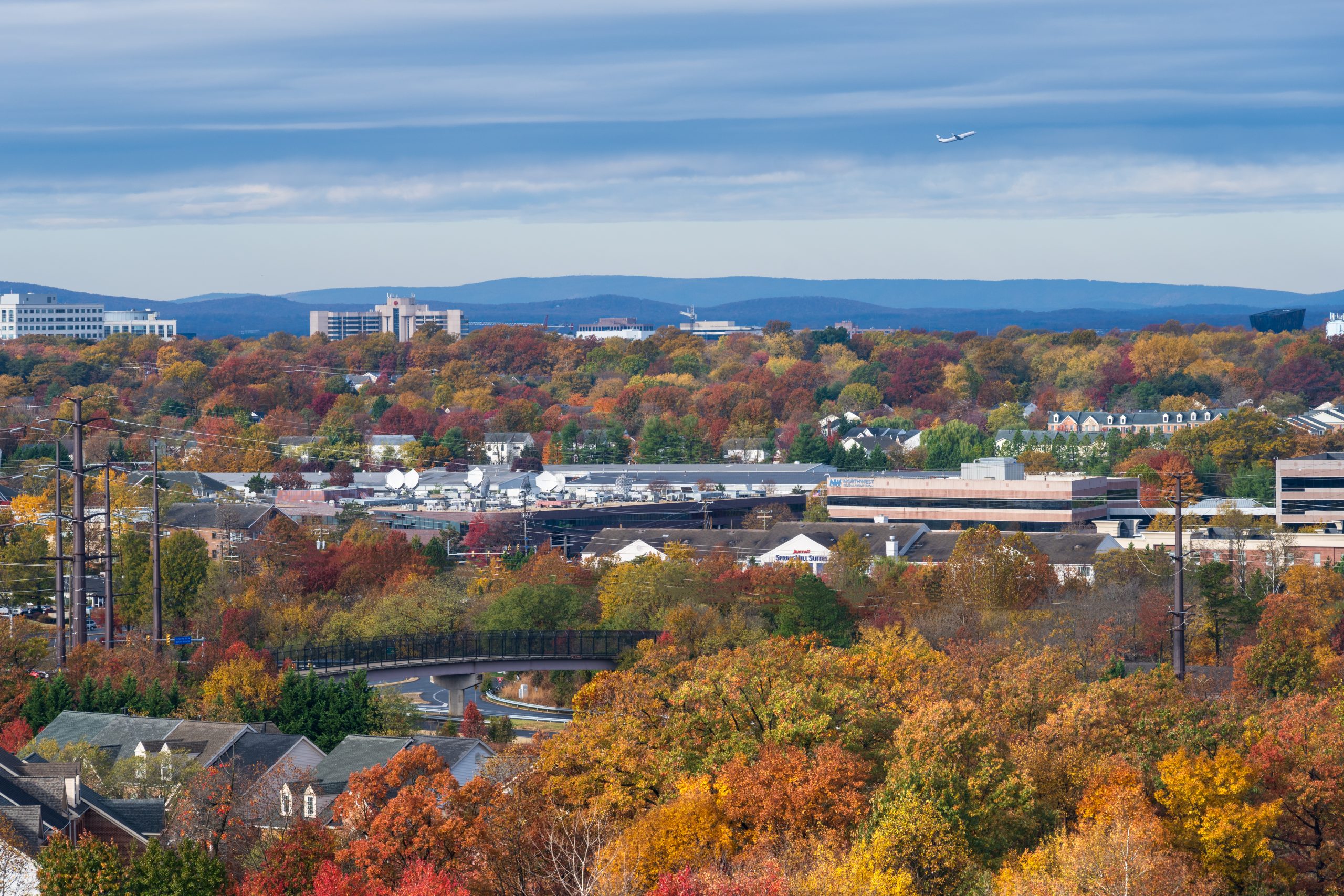 A landscape photo looks out over the lush scenery of Reston Virginia to the Blue Ridge Mountains as the seasons change; a plane in the sky is from Dulles Airport.