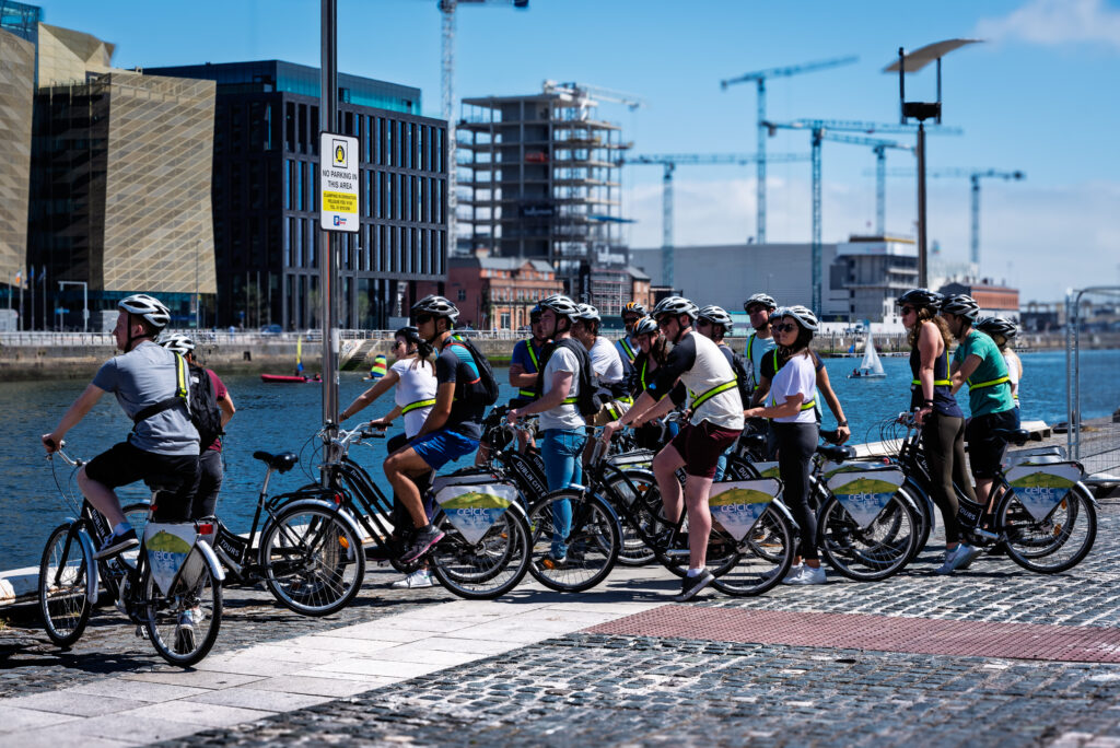 Group of helmeted cyclists on a guided bicycle tour beside the River Liffey waterfront in Dublin, with cranes and sailboats behind them.