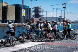 Group of helmeted cyclists on a guided bicycle tour beside the River Liffey waterfront in Dublin, with cranes and sailboats behind them.