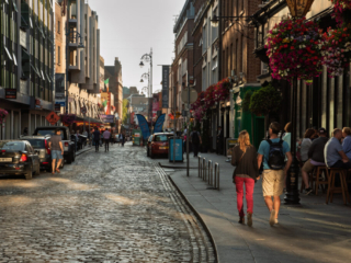 Busy cobblestone street in central Dublin lined with traditional pubs, colorful hanging flower baskets, pedestrians and parked cars in warm evening light.