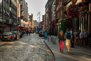 Busy cobblestone street in central Dublin lined with traditional pubs, colorful hanging flower baskets, pedestrians and parked cars in warm evening light.