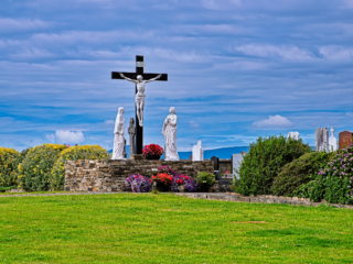 Calvary crucifix with white religious statues, colorful flowers and gravestones in a carefully maintained cemetery in rural Ireland.