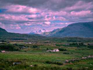 Rocky mountains rise above green fields, scattered farmhouses and stone walls in a broad rural valley in western Ireland beneath pink-tinted clouds.
