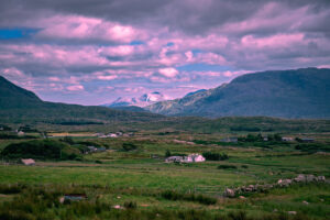 Rocky mountains rise above green fields, scattered farmhouses and stone walls in a broad rural valley in western Ireland beneath pink-tinted clouds.