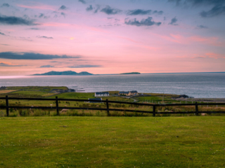 Pink sunset over Ireland's Atlantic coast with offshore islands, coastal homes, green pasture and a dark wooden fence in the foreground.