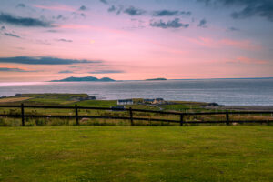 Pink sunset over Ireland's Atlantic coast with offshore islands, coastal homes, green pasture and a dark wooden fence in the foreground.