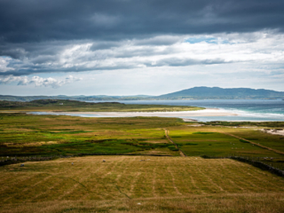 Wide view of Ireland's Atlantic coast with green fields, a white sand beach, blue water and distant mountains beneath storm clouds.