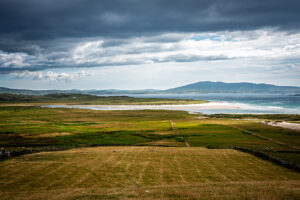 Wide view of Ireland's Atlantic coast with green fields, a white sand beach, blue water and distant mountains beneath storm clouds.