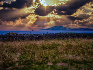 Golden sunrays break through dark clouds over the Atlantic Ocean and a mountain on Ireland's coast, viewed beyond a stone wall and meadow.
