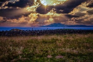 Golden sunrays break through dark clouds over the Atlantic Ocean and a mountain on Ireland's coast, viewed beyond a stone wall and meadow.