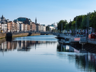 River Liffey cityscape in Dublin with the Ha'penny Bridge, historic quayside buildings, riverside trees and reflections on calm water.