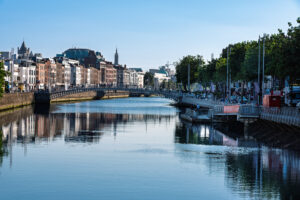River Liffey cityscape in Dublin with the Ha'penny Bridge, historic quayside buildings, riverside trees and reflections on calm water.