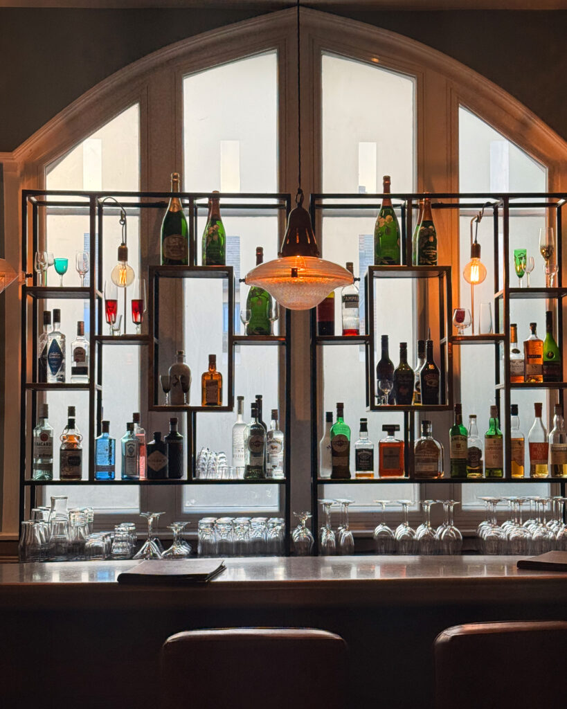 Bottles and rows of glasses fill black bar shelves beneath an arched window and glowing pendant lights.
