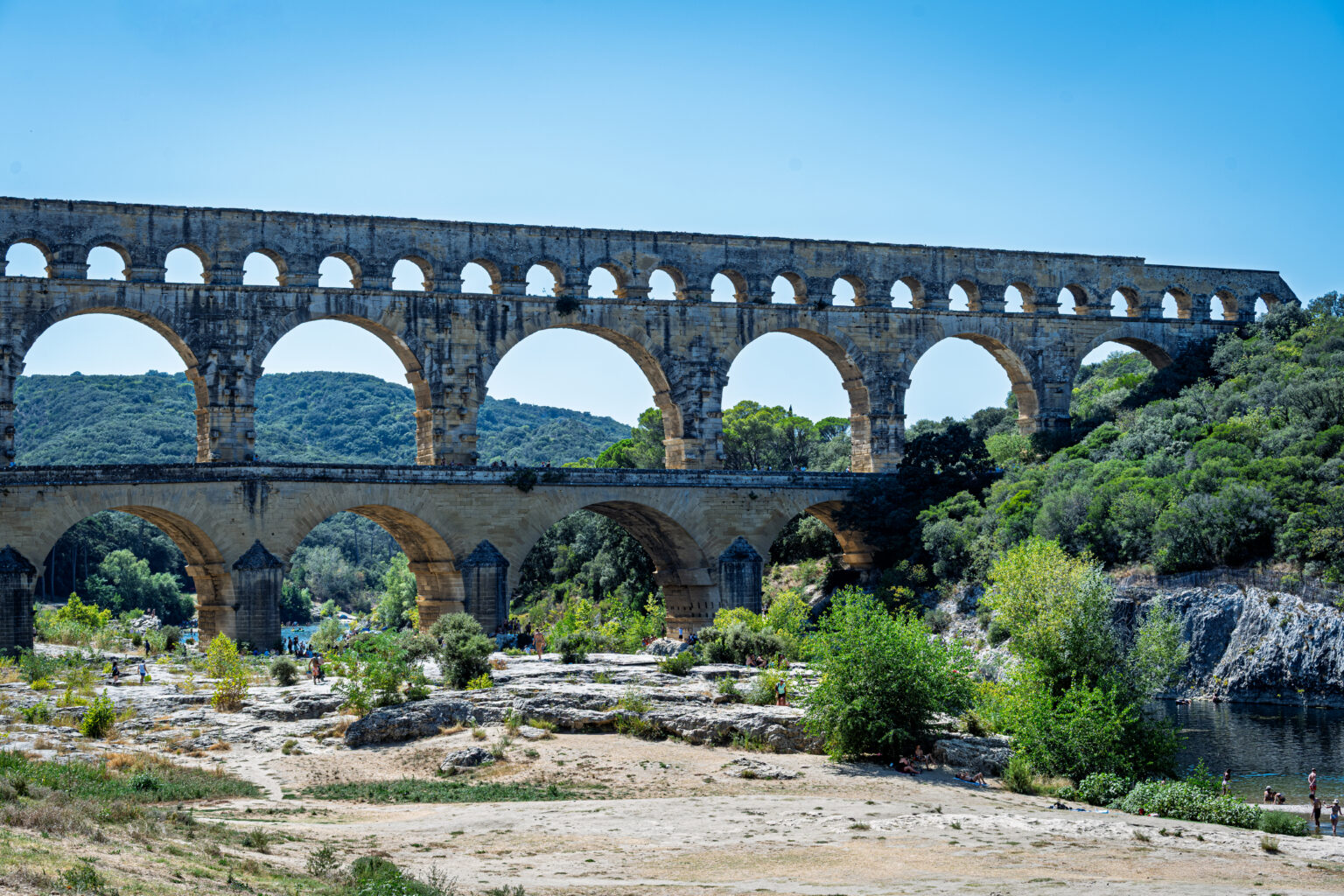 The stacked arches of the Pont du Gard rise above a rocky riverbank and green vegetation in southern France.