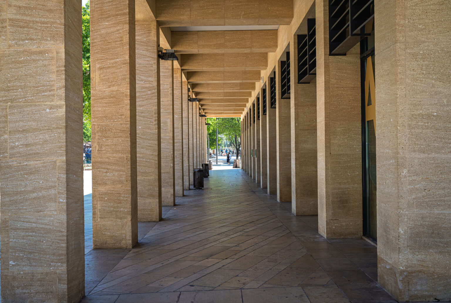 Rectangular columns and overhead beams form a long shaded arcade leading toward daylight.
