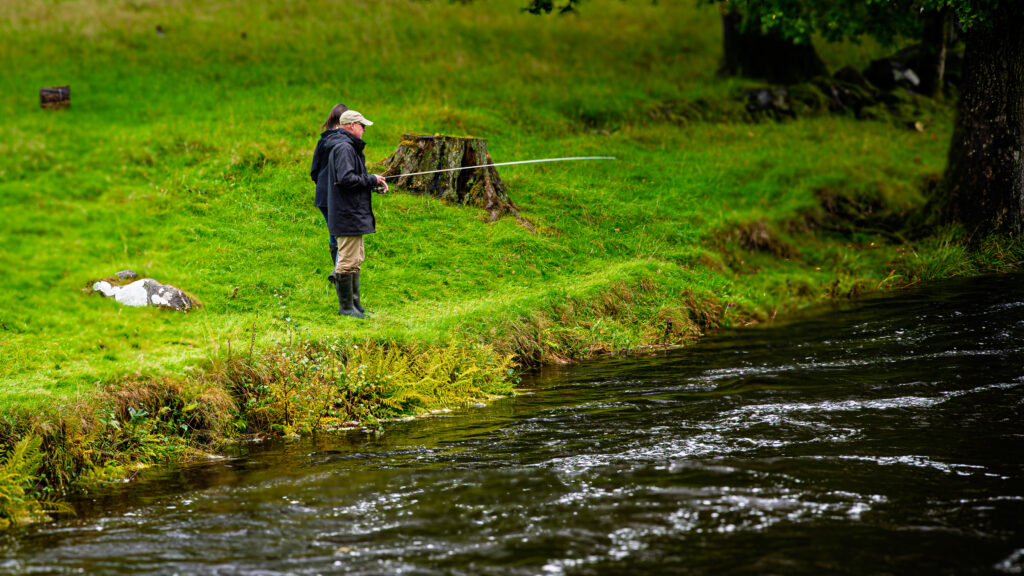 An angler with a fishing rod and a second person beside a rippling river under leafy trees in Eryri, Wales.