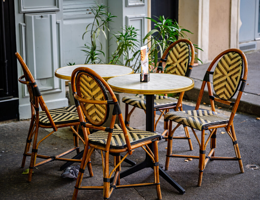 Woven bistro chairs surround a small round table outside a café with pale doors and potted plants.