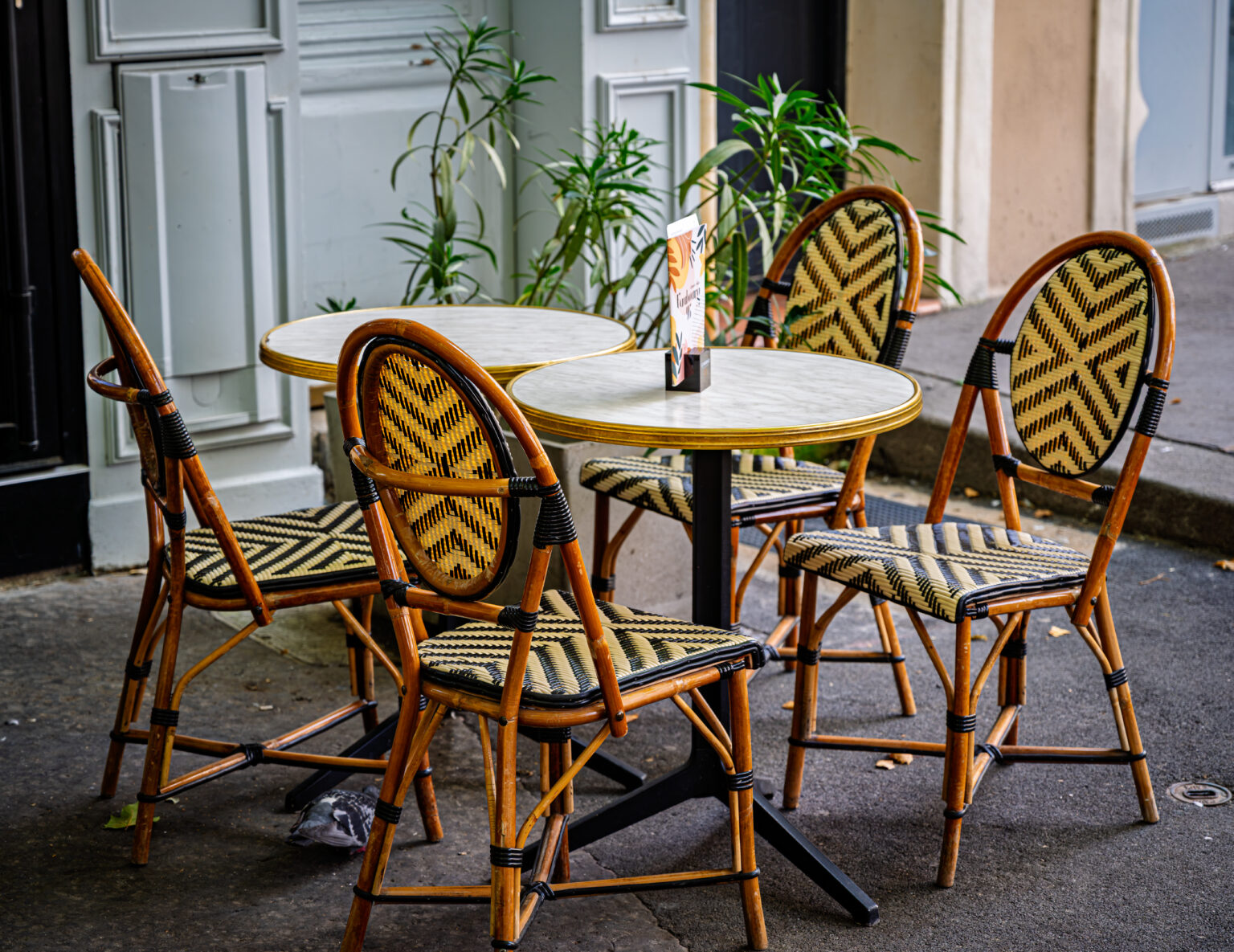 Woven bistro chairs surround a small round table outside a caf&eacute; with pale doors and potted plants.