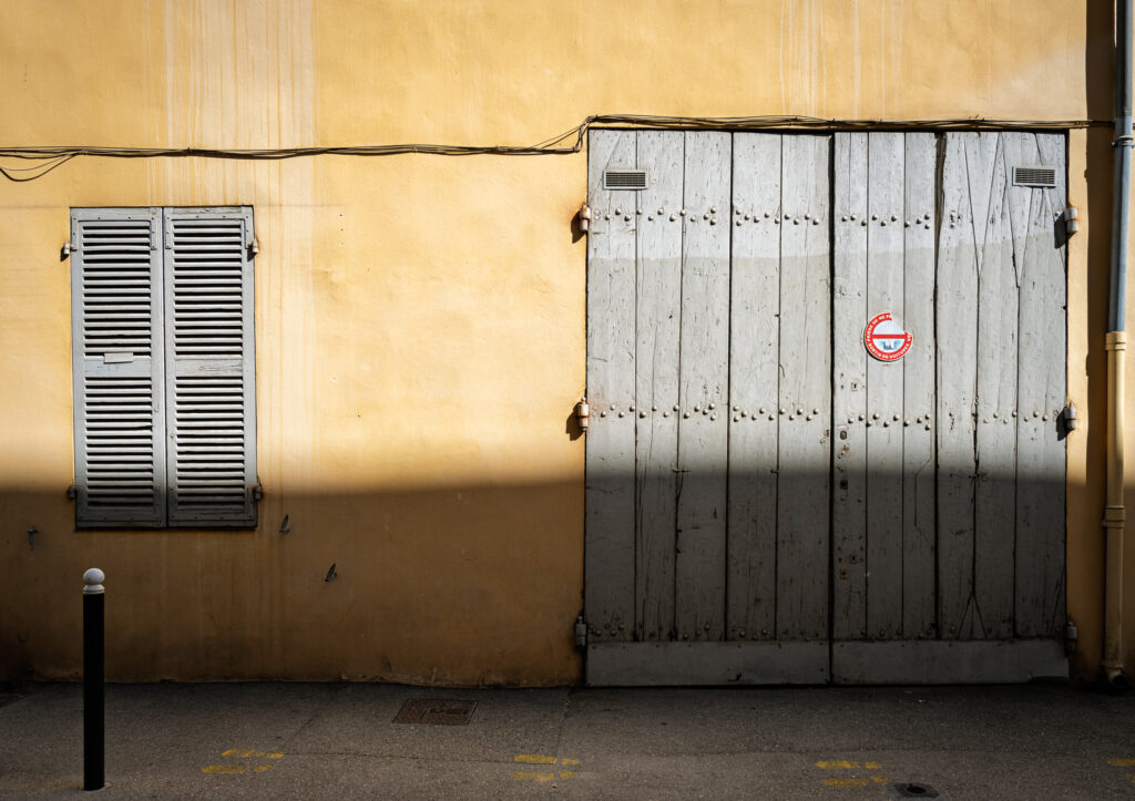 A yellow wall in Provence frames closed gray shutters and a broad wooden door, with a horizontal shadow crossing the façade.