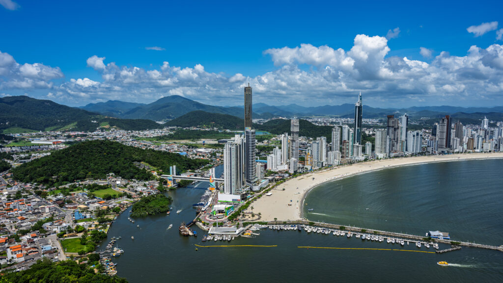 A broad elevated view shows a curving beach, dense high-rise skyline, river mouth and green hills in Balneário Camboriú.