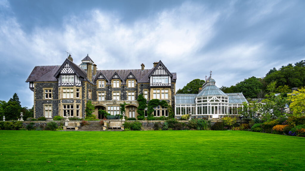 Bodnant Hall&rsquo;s gabled fa&ccedil;ade and glass conservatory face a broad green lawn in Wales.