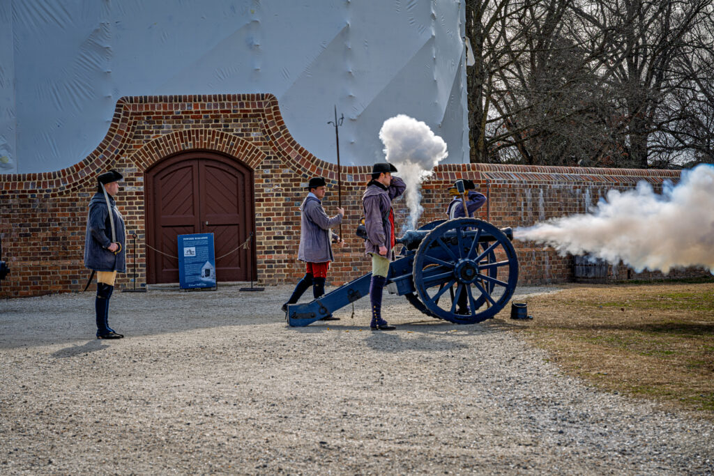 Costumed interpreters fire a cannon beside a brick wall at Colonial Williamsburg as white smoke billows across the courtyard.