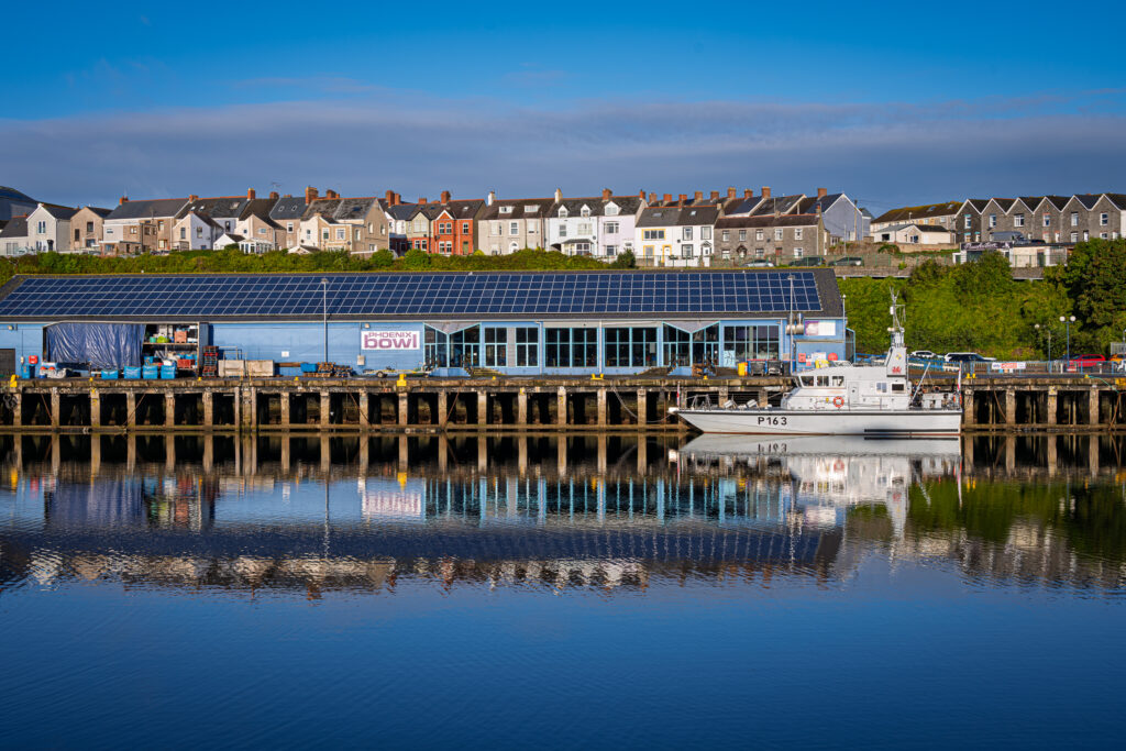 A white P163 vessel and the solar-roofed Phoenix Bowl building reflect in Milford Haven dock water.