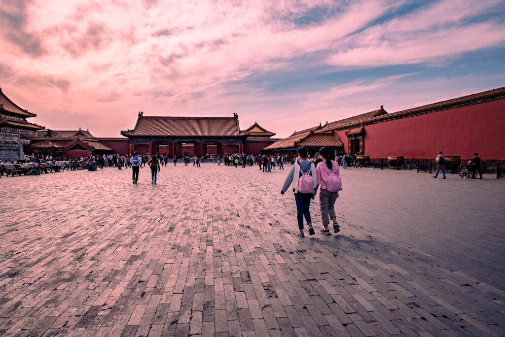 Visitors cross a broad stone courtyard in Beijing’s Forbidden City beneath a pink-toned sky.