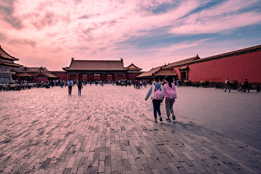 Visitors cross a broad stone courtyard in Beijing&rsquo;s Forbidden City beneath a pink-toned sky.