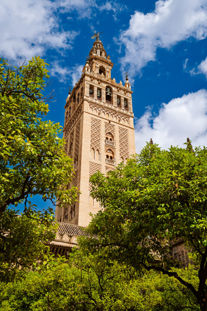 The Giralda rises above green trees beneath blue sky and white clouds in Seville.