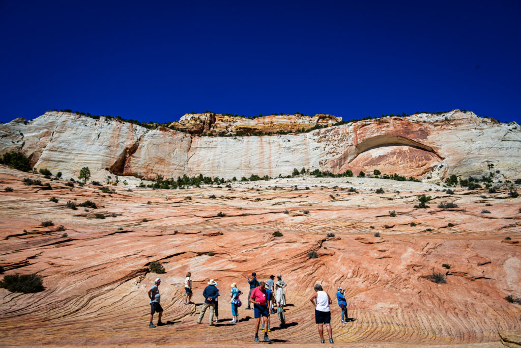 A group of hikers stands on rippled sandstone below a broad pale cliff and shallow alcove.