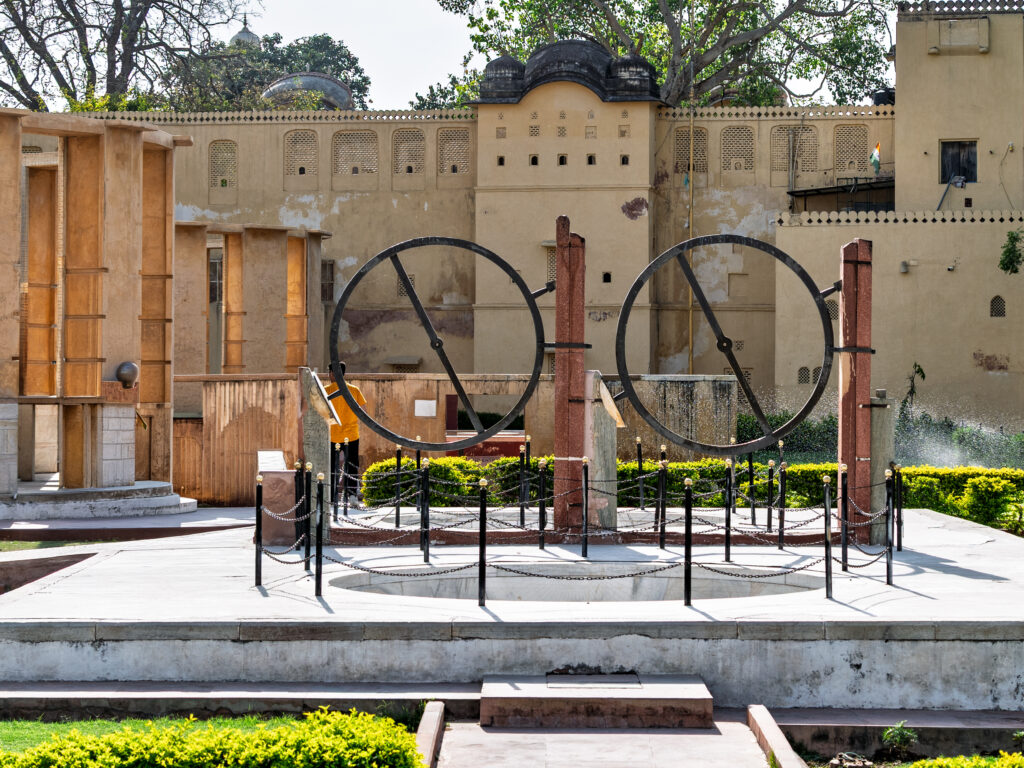 Two circular astronomical instruments stand on a platform in front of a weathered ochre wall.