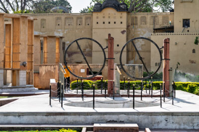 Two circular astronomical instruments stand on a platform in front of a weathered ochre wall.