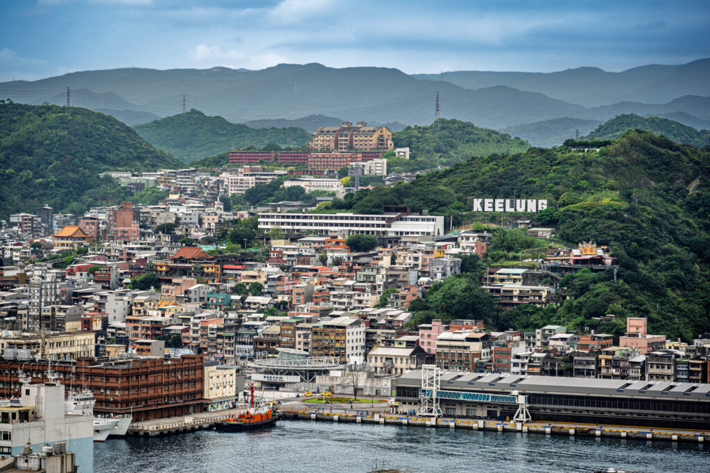 Dense waterfront buildings fill Keelung beneath green hills and a prominent city sign.