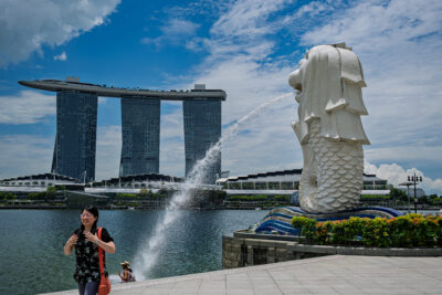 A pedestrian crosses the waterfront plaza near the Merlion with Marina Bay Sands in the background.