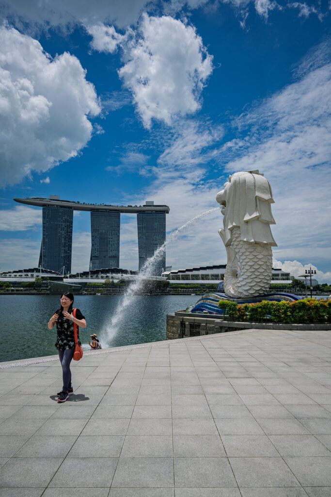 A pedestrian crosses the waterfront plaza near the Merlion with Marina Bay Sands in the background.