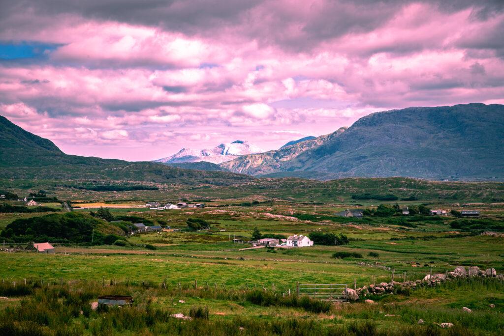 Rocky mountains rise above green fields, scattered farmhouses and stone walls in a broad rural valley in western Ireland beneath pink-tinted clouds.