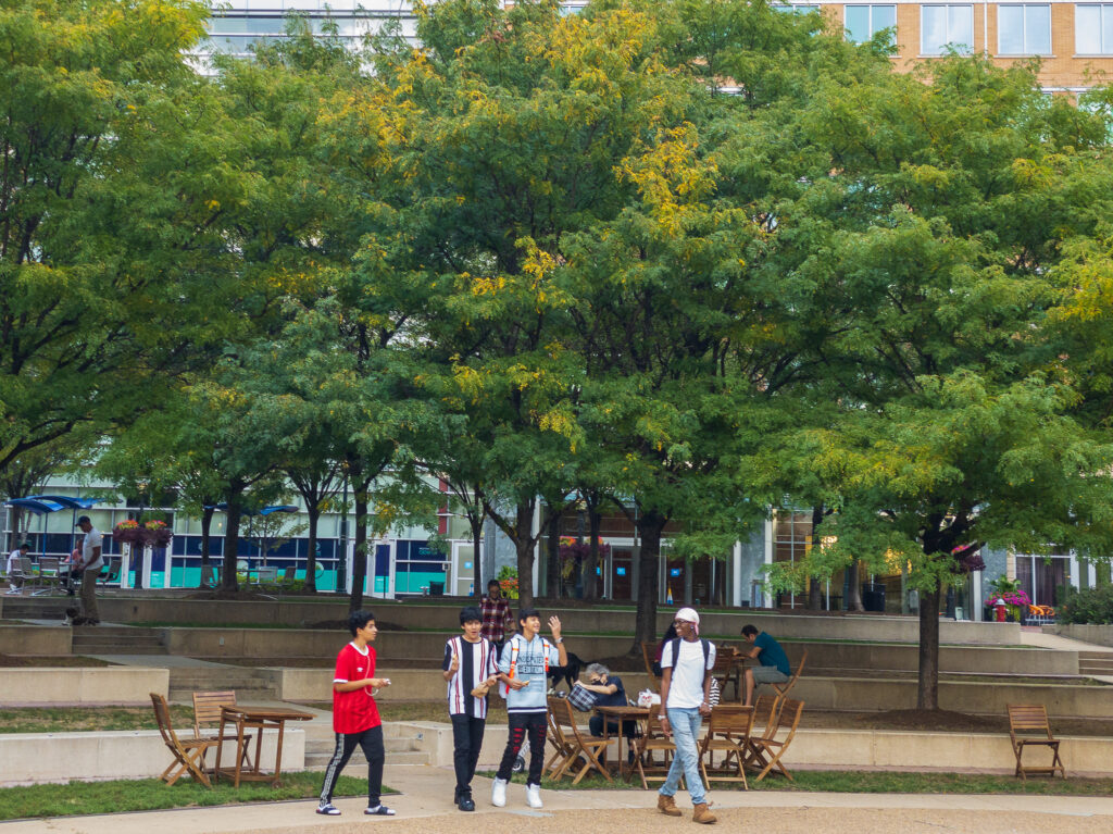 A group walks past wooden tables and chairs beneath green trees in Reston Town Center Park, Virginia.