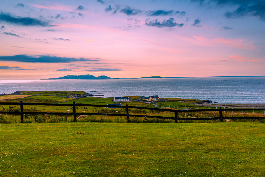 Pink sunset over Ireland's Atlantic coast with offshore islands, coastal homes, green pasture and a dark wooden fence in the foreground.