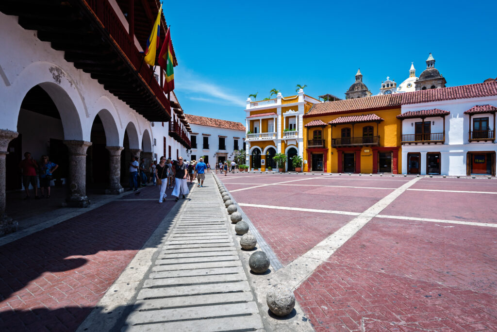 Shaded arcades beside sunlit Plaza de la Aduana in Cartagena, with colorful facades and church domes.