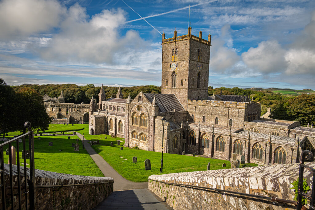 St Davids Cathedral’s square tower rises above stone roofs, green lawns and gravestones in Wales.