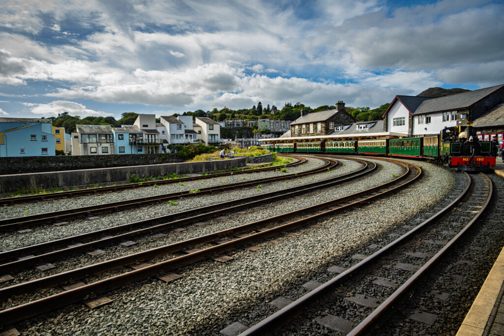 Curving railway tracks lead to a green steam train at Porthmadog Harbour Station.