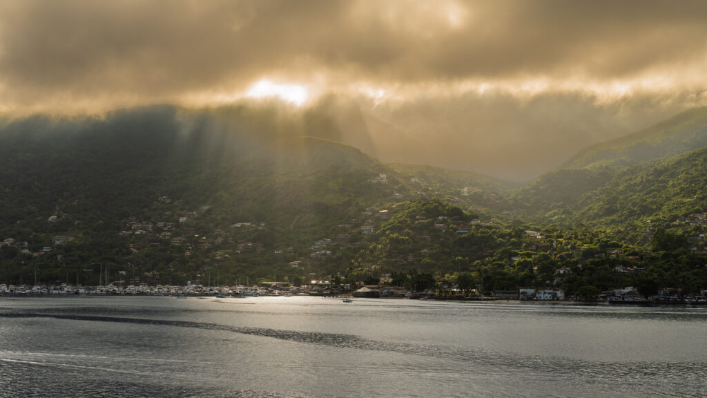 Sunbeams break through heavy clouds over forested coastal hills and water in Ilhabela.
