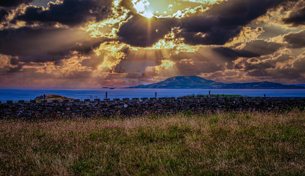 Golden sunrays break through dark clouds over the Atlantic Ocean and a mountain on Ireland's coast, viewed beyond a stone wall and meadow.