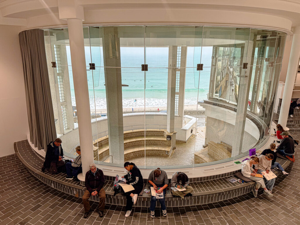 Curved white interior of Tate St Ives with visitors and turquoise sea visible through large windows.