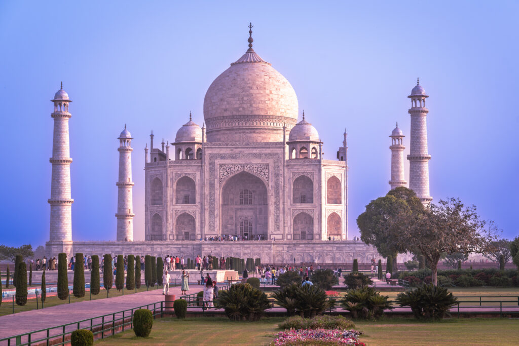 The ivory-colored Taj Mahal rises above garden beds and a visitor walkway beneath a pastel sky.