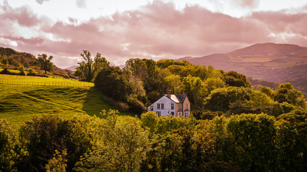 A white cottage sits among green fields and trees with distant hills beneath a pink-toned sky.