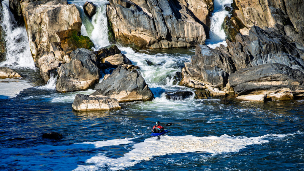 A lone kayaker crosses blue water and white foam below rugged rocks and cascading waterfalls.