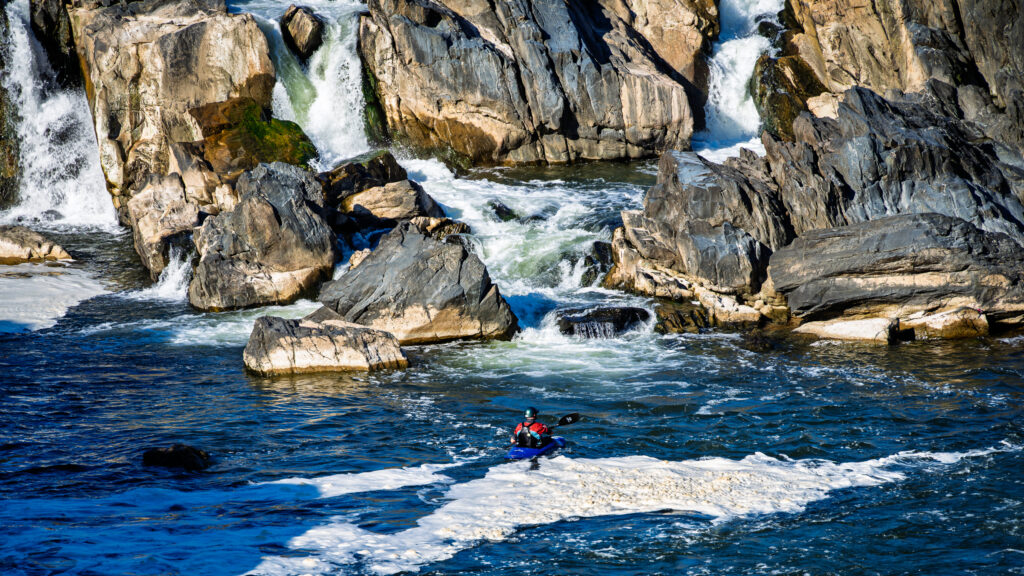 A lone kayaker crosses blue water and white foam below rugged rocks and cascading waterfalls.