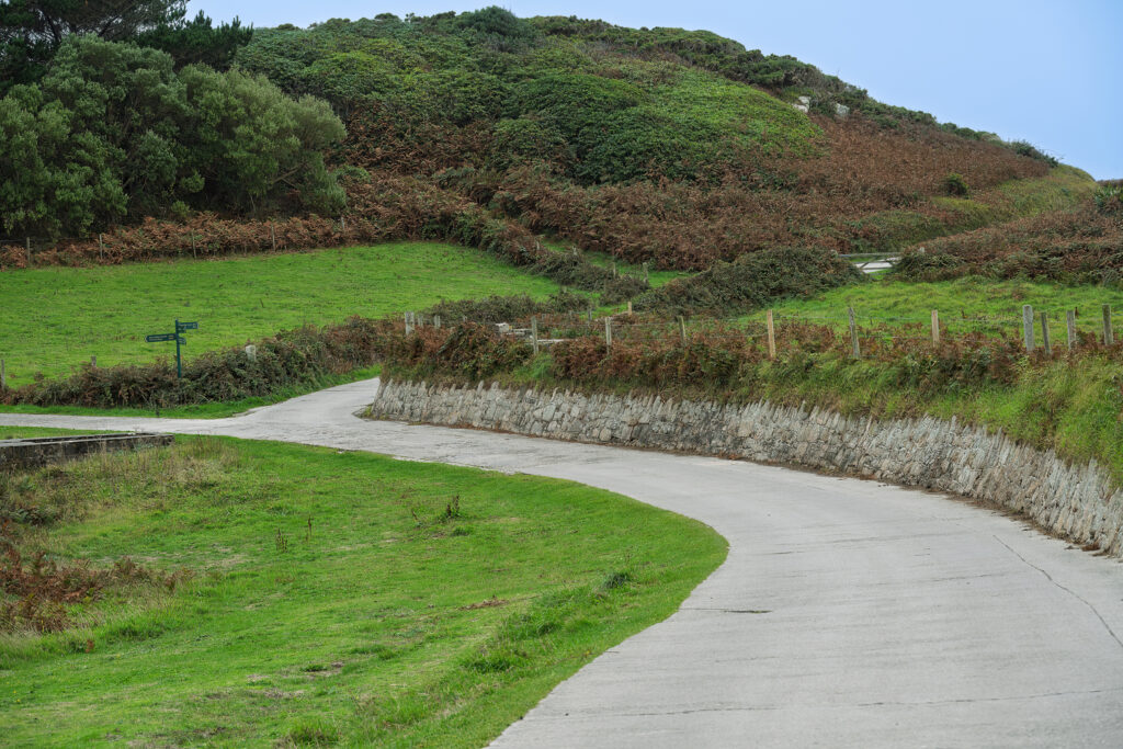 Winding paved coastal path through green vegetation on Tresco in the Isles of Scilly beneath a moody sky.